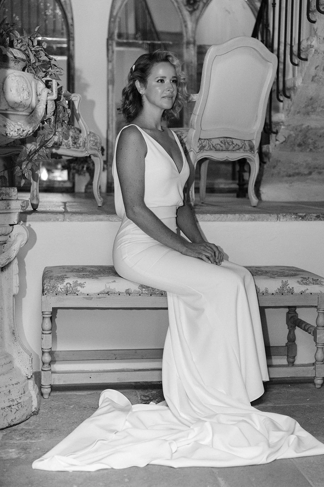 A woman in a white dress sitting on a vintage bench indoors, with a wedding dress trailing on the floor in front of her, in a room decorated with antique furniture and plants.
