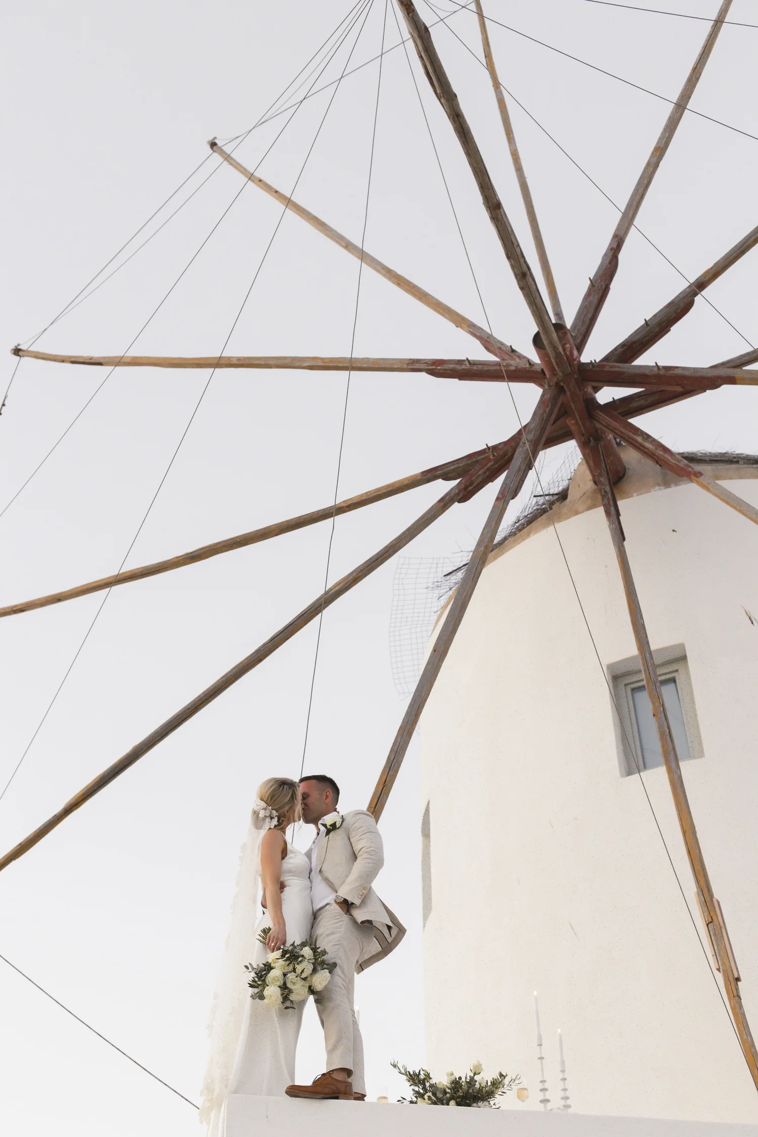 A bride and groom sharing a kiss during their wedding ceremony in front of a white church, under a wooden structure with overhead wires and candles on the ground.