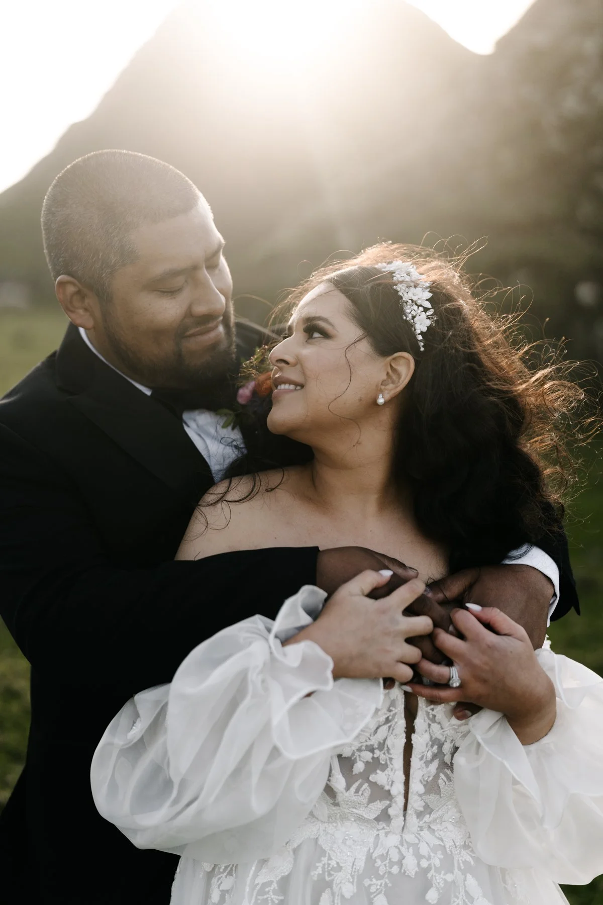 A newlywed couple embracing outdoors during sunset, with the groom in a black tuxedo and the bride in a white lace wedding dress, smiling at each other.