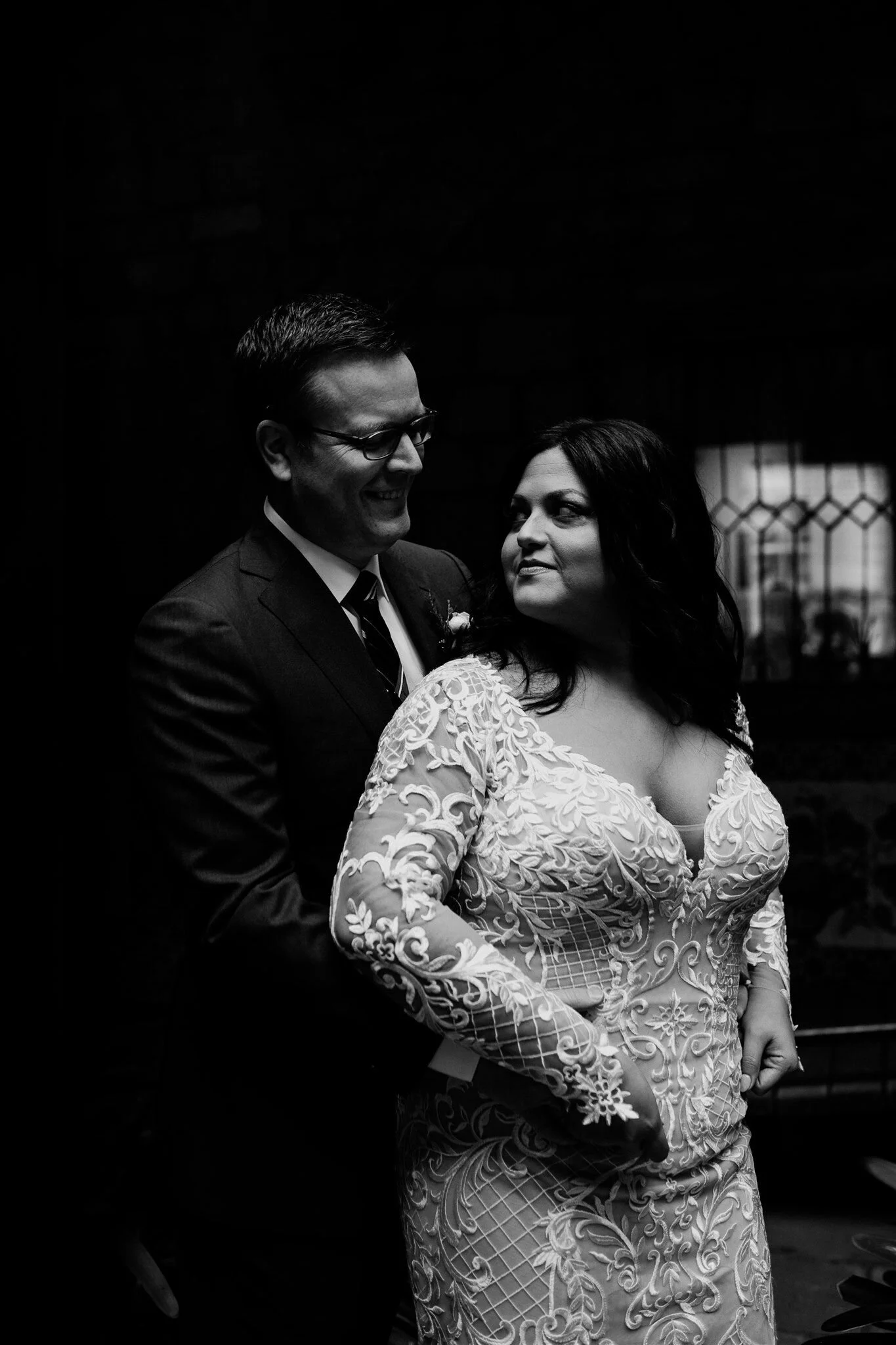 Black and white photo of a couple dressed in wedding attire, smiling and looking at each other, standing indoors with a dark background.