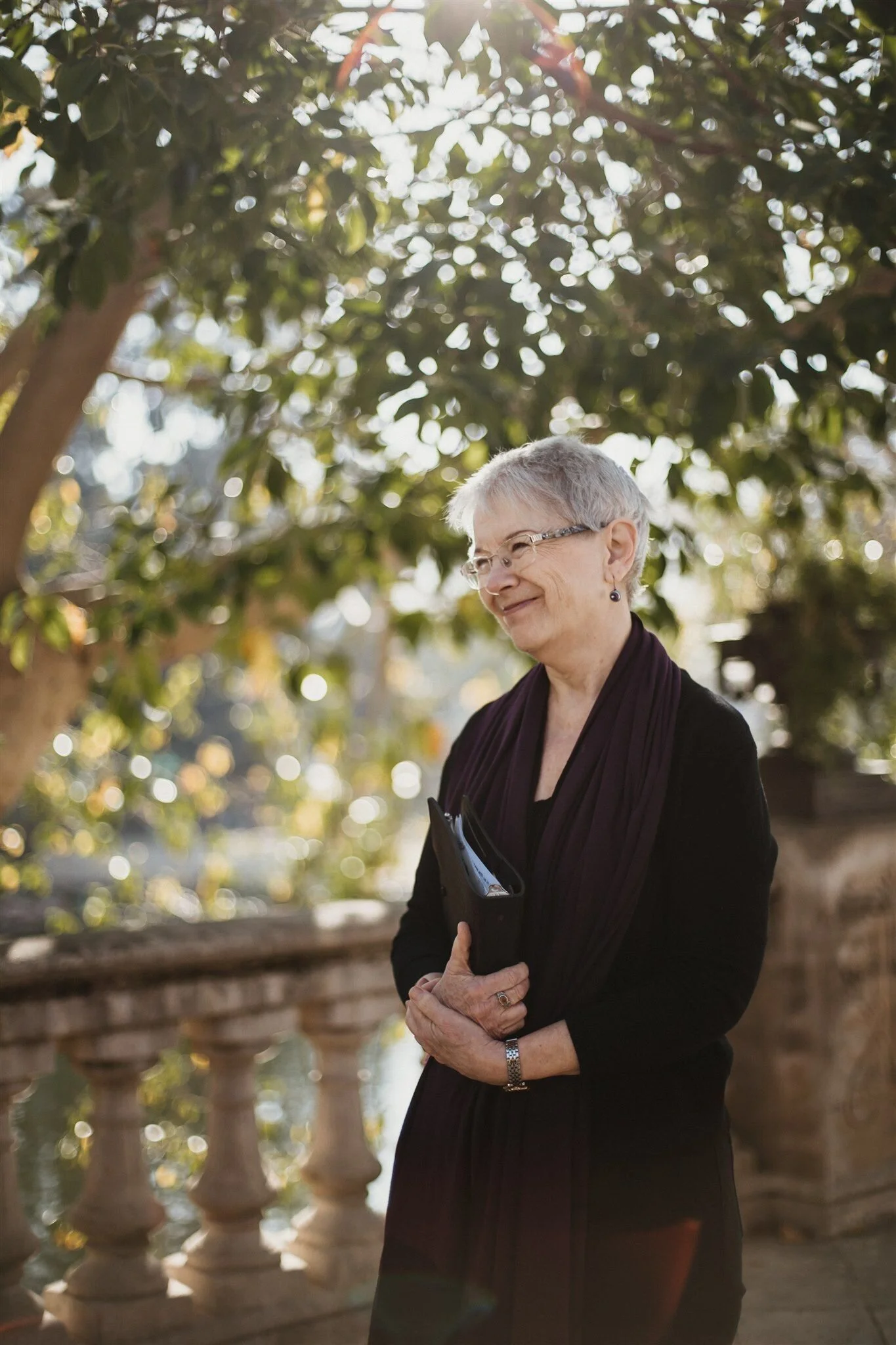 An elderly woman standing outdoors in front of a large leafy tree with sunlight filtering through the branches. She has short gray hair, glasses, and is dressed in dark clothing, holding something in her hands and smiling softly.