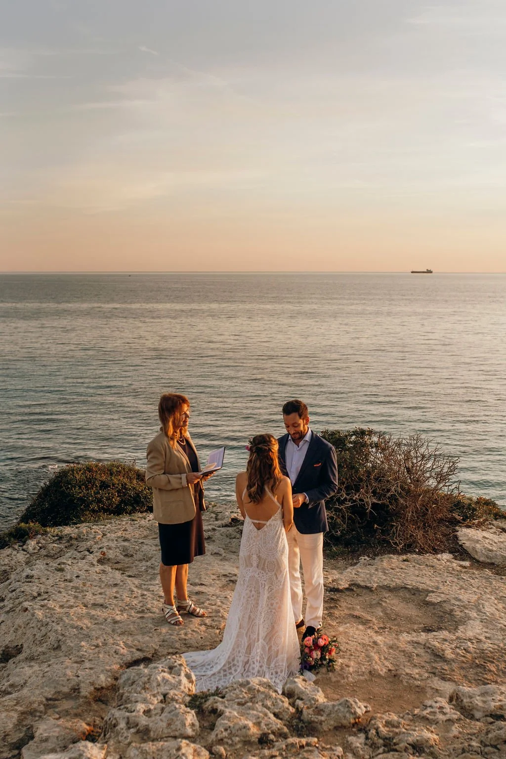 A couple getting married on a rocky beach at sunset, with an officiant standing nearby reading from a book, and the ocean and a distant ship in the background.