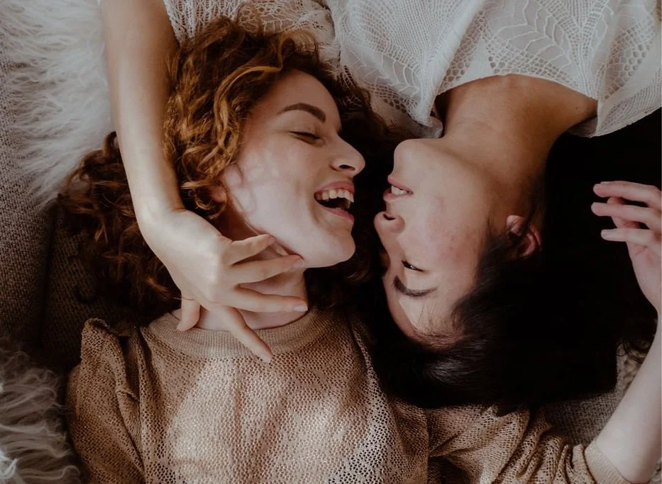Two women lying on a bed, smiling and looking at each other, sharing a moment of affection.