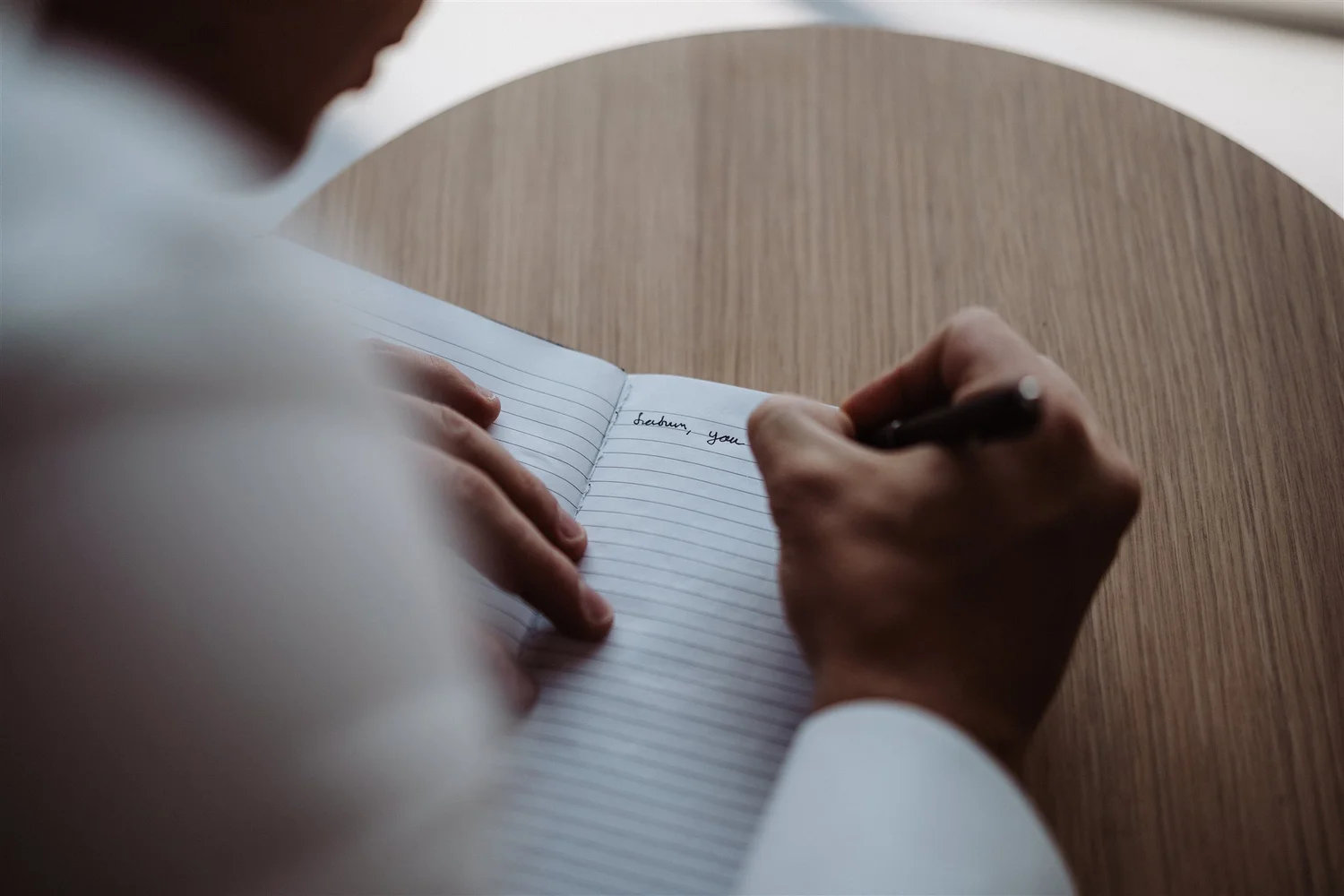 A person in a suit writing in a lined notebook at a wooden table.