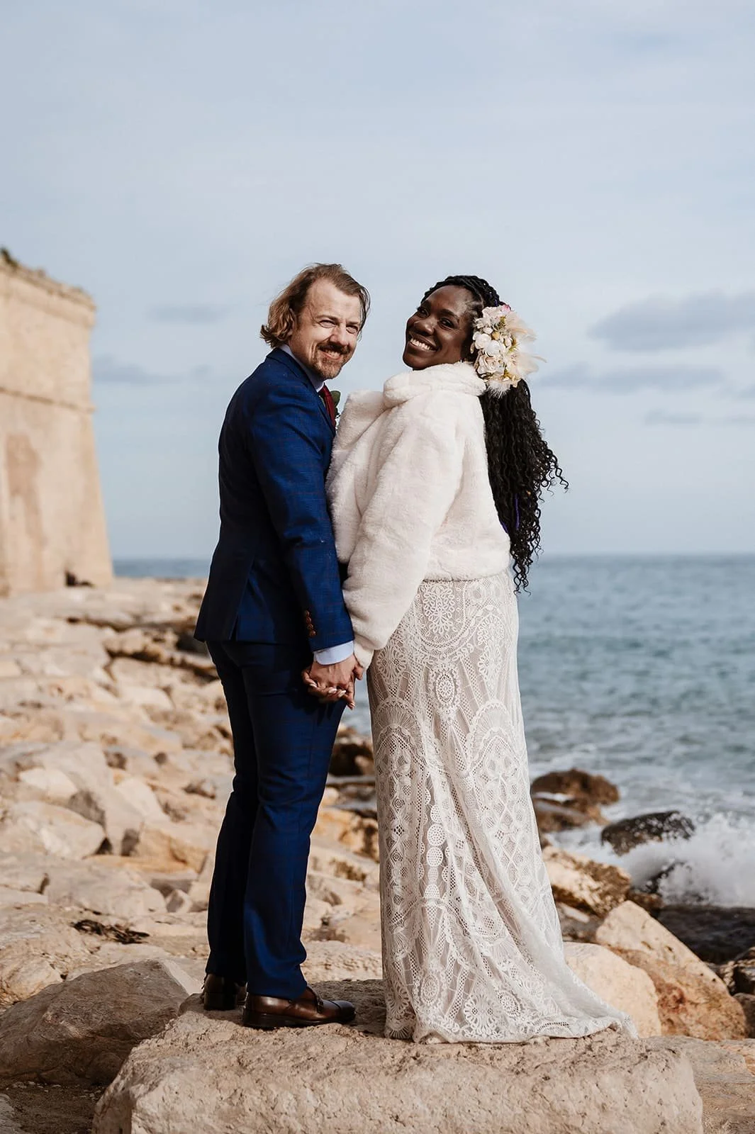 A happy couple standing on a rocky beach holding hands, with the sea and cloudy sky in the background. The woman has long curly hair with flowers and wears a white dress and fuzzy coat, the man has wavy hair with a beard and wears a blue suit.
