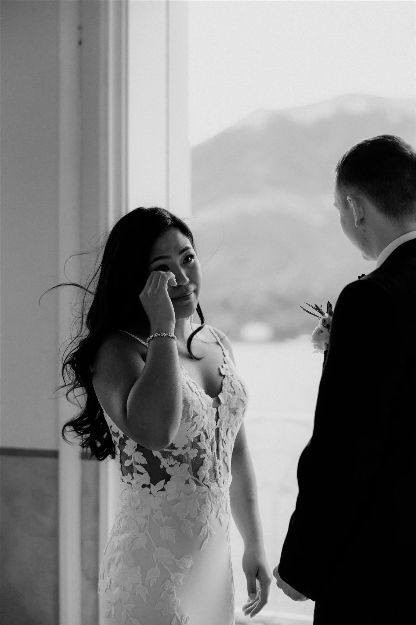 Black and white photo of a woman in a lace wedding dress standing in front of a man in a tuxedo, holding hands, with mountains visible through a window in the background.