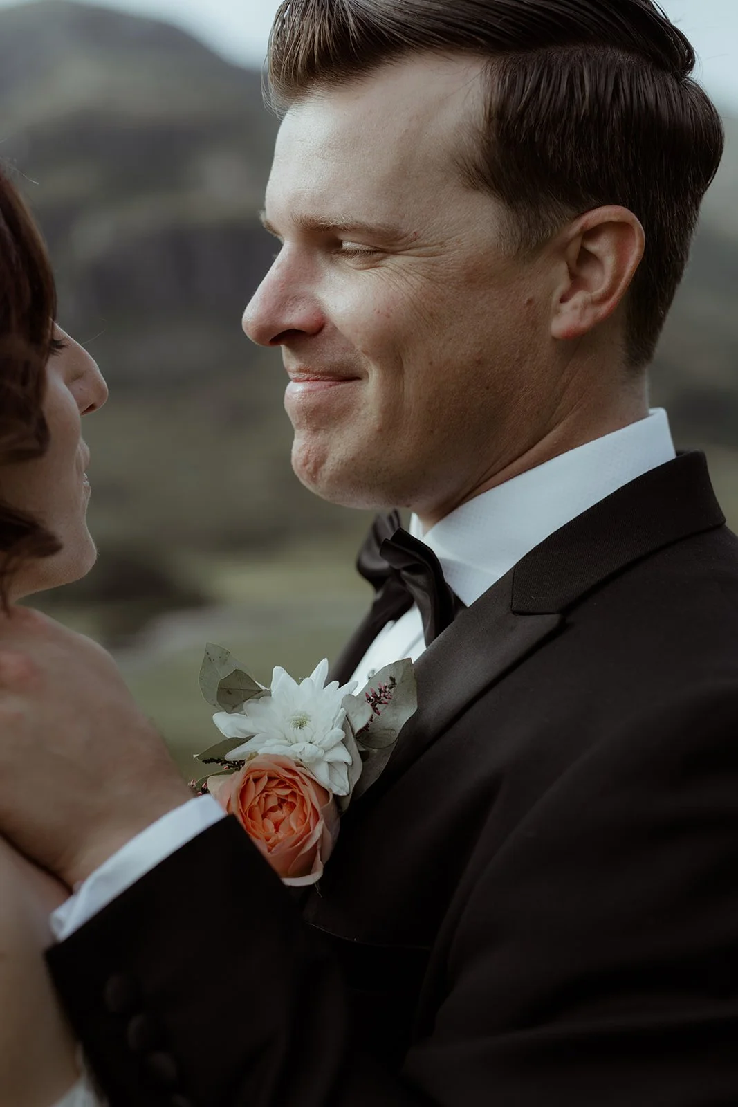 A close-up of a bride and groom smiling at each other outdoors with a blurred natural landscape background. The groom wears a black tuxedo with a white shirt and a black bowtie, and has a boutonniere with white and peach flowers.