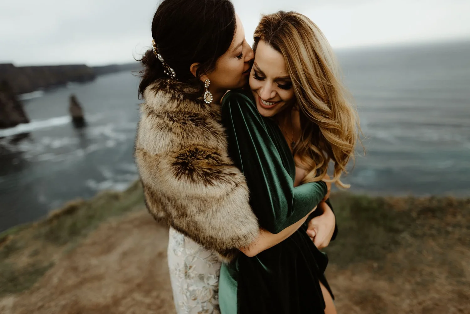 Two women embrace by the ocean on a rocky coastline, with overcast skies.