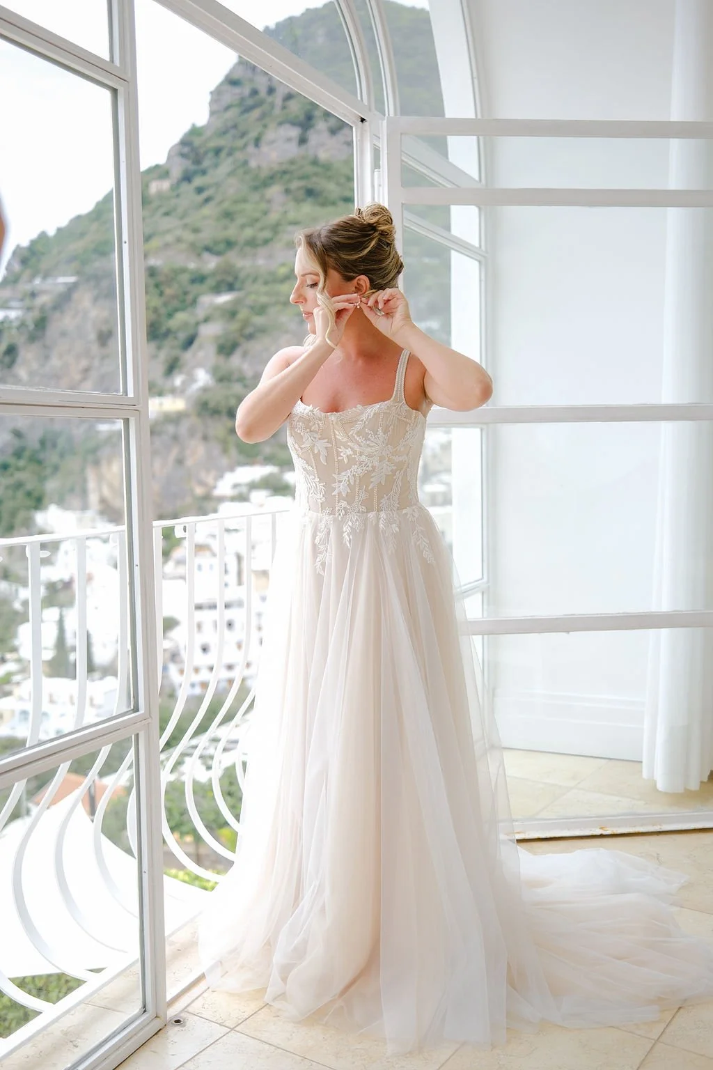 A woman in a wedding dress standing near large windows, adjusting her earring, with a mountain landscape in the background.