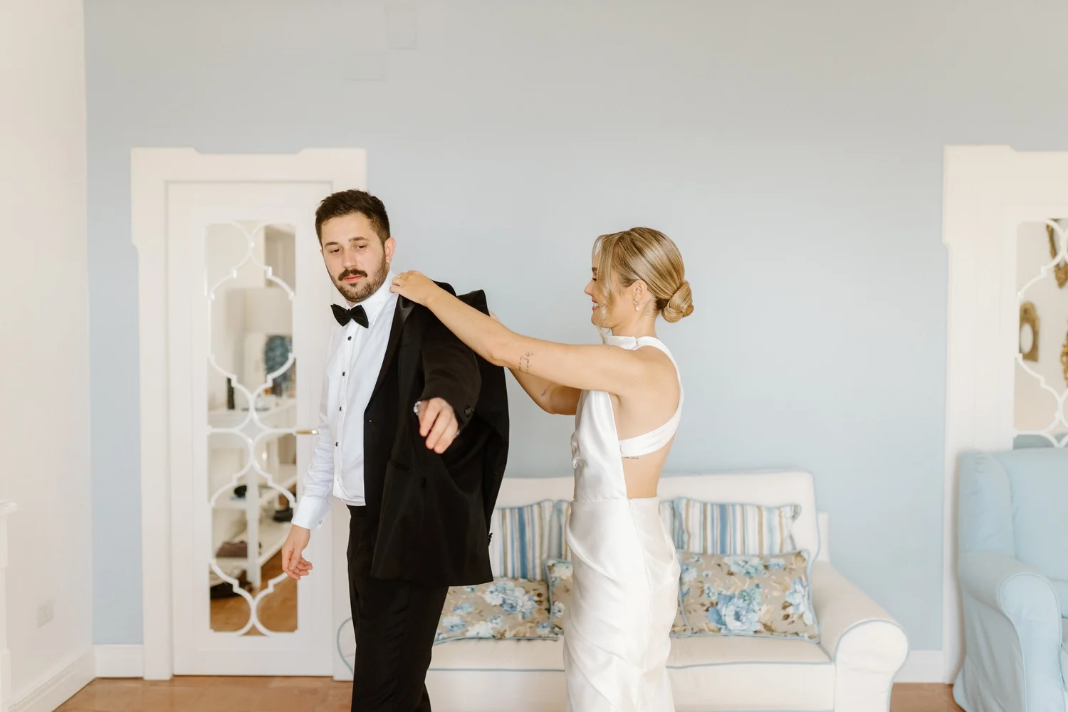 A woman in a white dress helping a man in a tuxedo with his jacket in a bright room with a decorative white door and a bed with a floral cover in the background.
