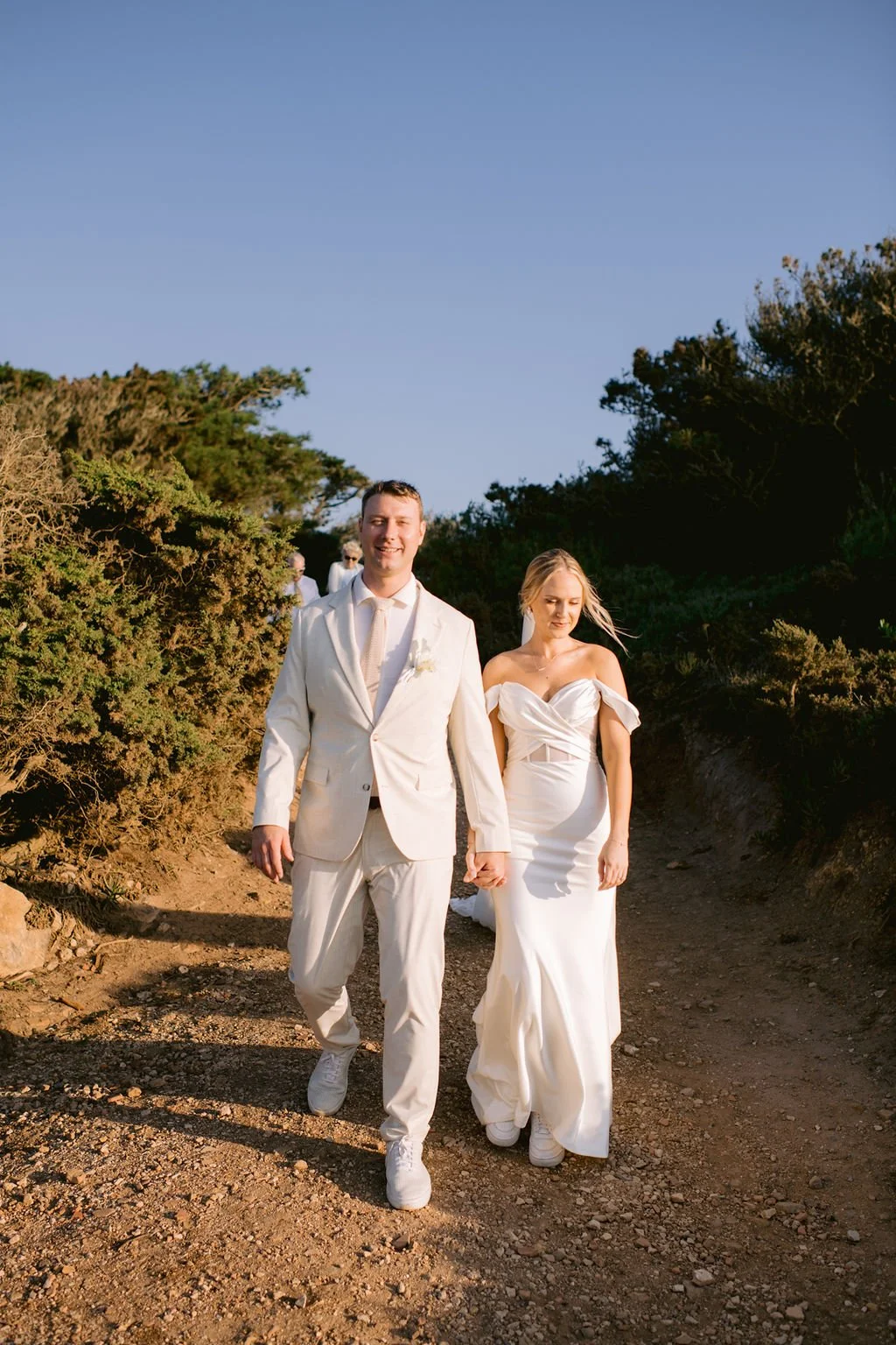 A newlywed couple walking hand in hand outdoors on a dirt trail, with greenery on either side, under a clear blue sky; the groom is wearing an off-white suit, and the bride is in a strapless white wedding dress.