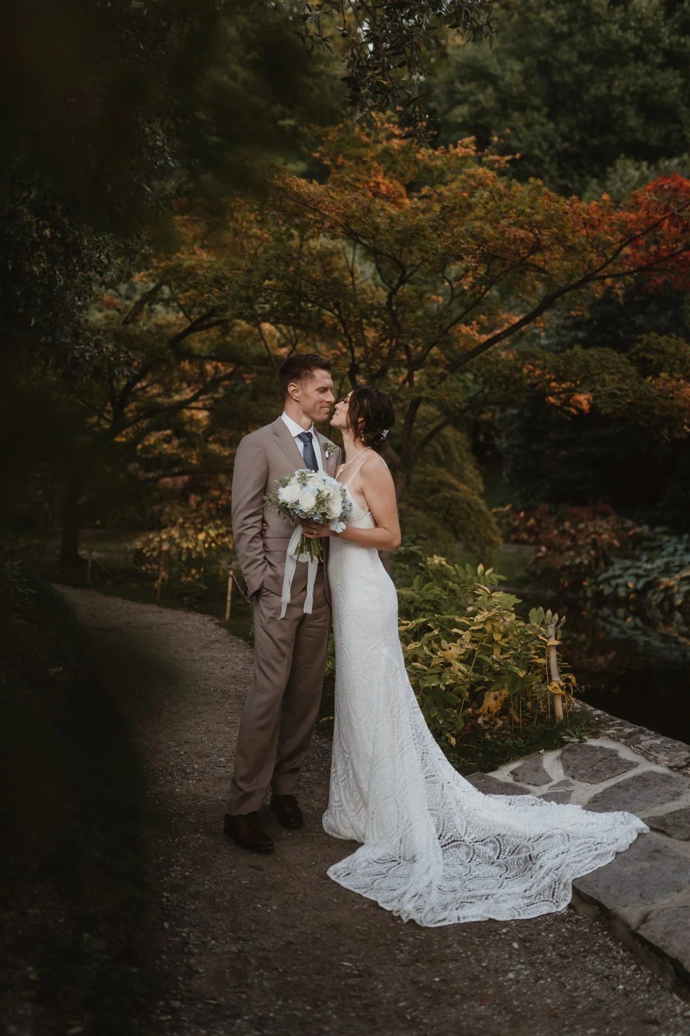 A bride and groom sharing a kiss in a park during autumn, surrounded by colorful trees and foliage.