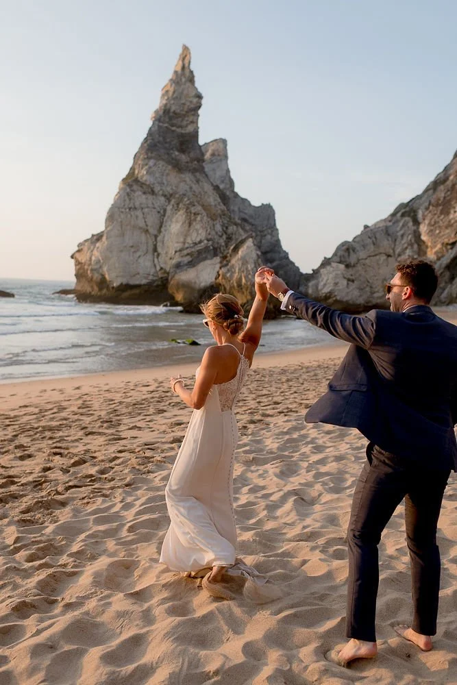 A couple in formal attire dancing barefoot on a sandy beach at sunset with large rock formations in the background.