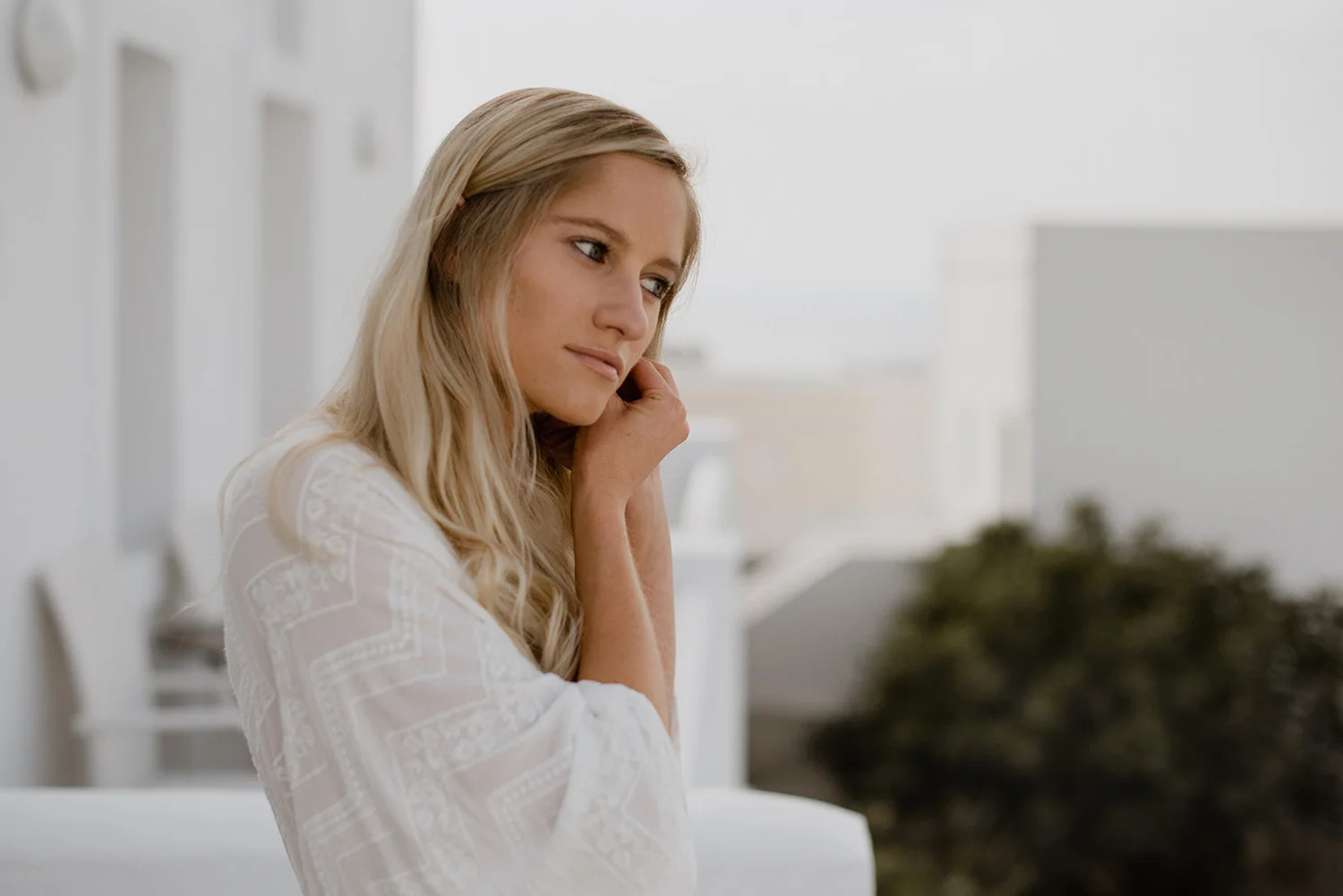 A woman with blonde hair and a white lace patterned top sitting indoors with her chin resting on her hand.