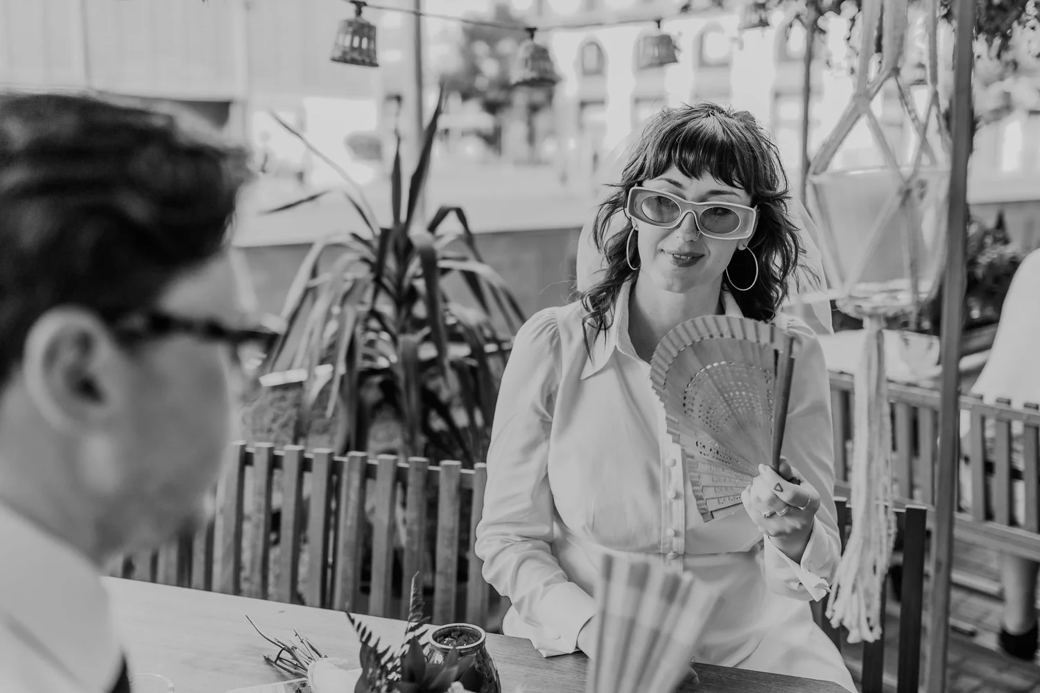 A woman with wavy hair, wearing sunglasses and hoop earrings, holding a fan and sitting at a table indoors. In the foreground, part of a child’s head is visible. The background features large windows and shelves filled with plants and decorative item