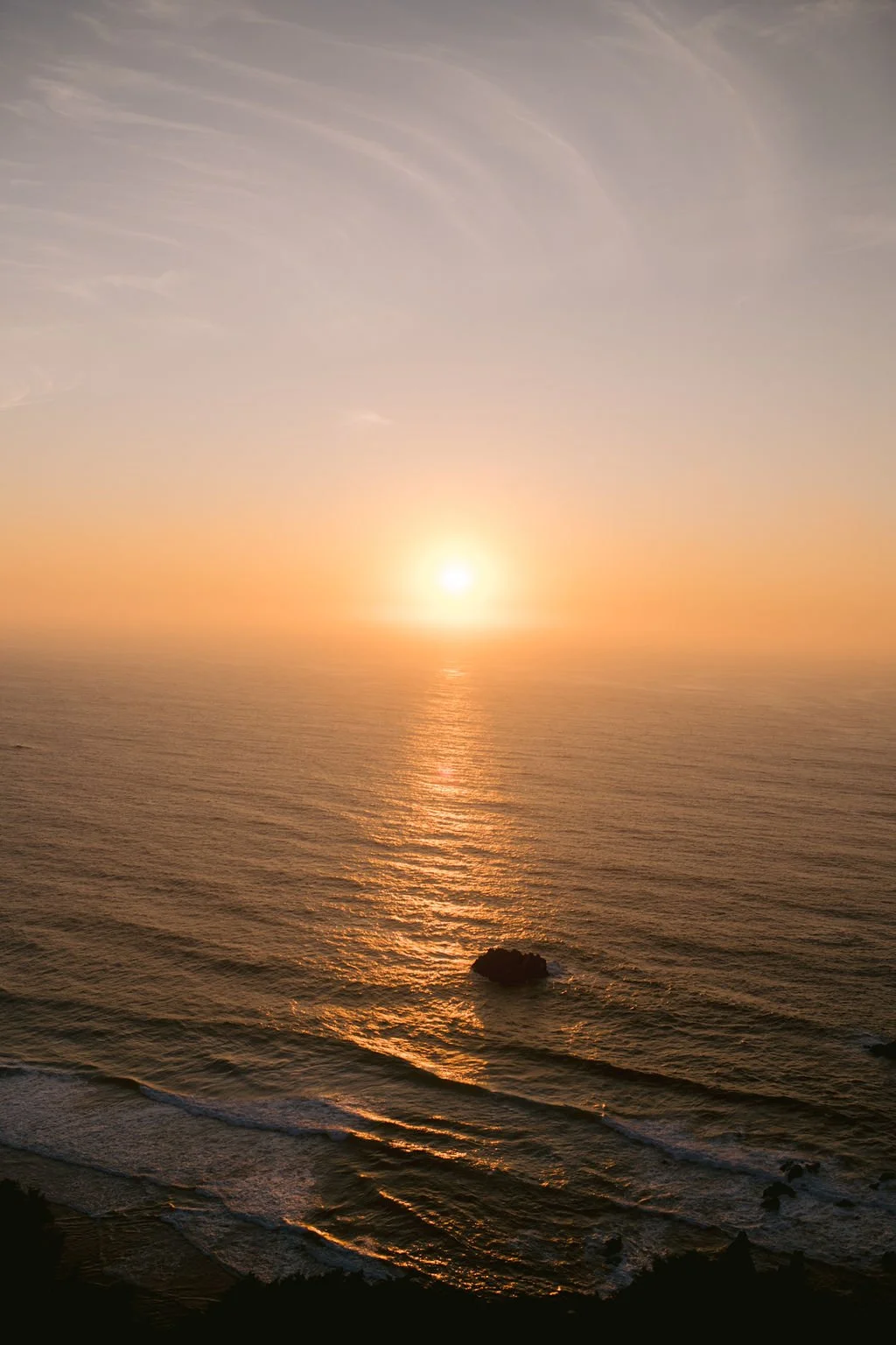 Sunset over the ocean with calm waves and a rock in the water.