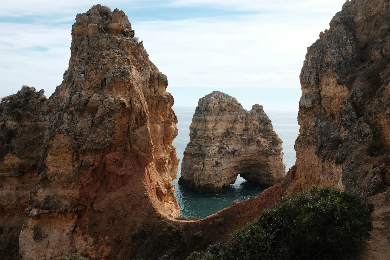 Rock formations along a coastline with an arch-shaped opening in the middle, overlooking the ocean under a partly cloudy sky.