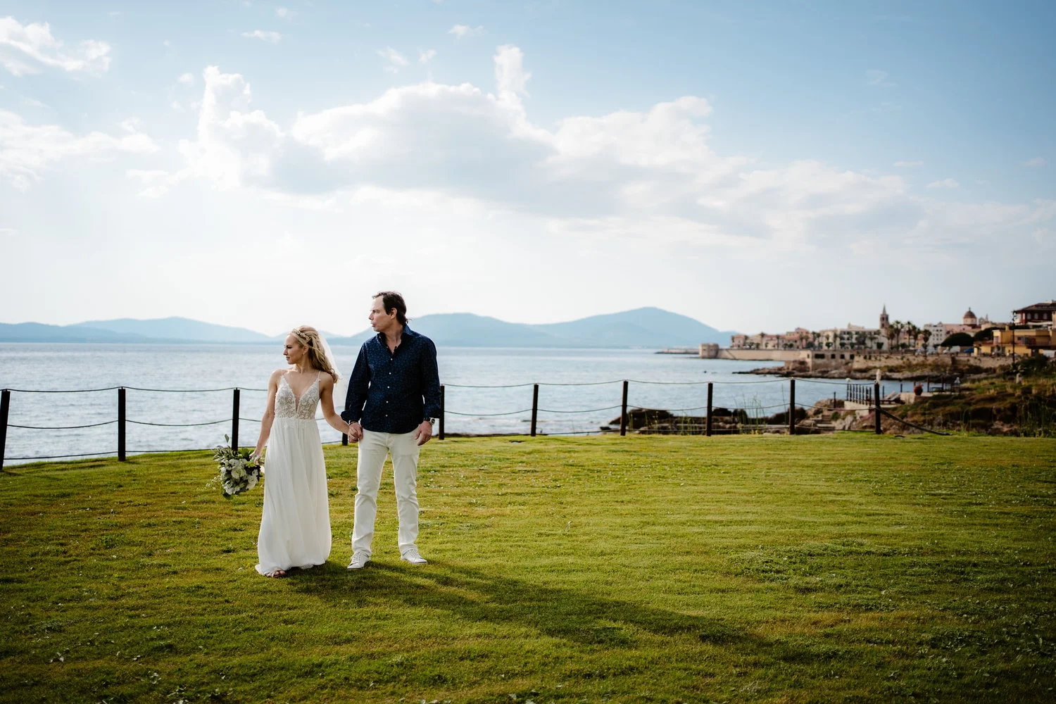 A bride and groom holding hands walk on a grassy area near a waterfront on a partly cloudy day, with a town and mountains in the background.