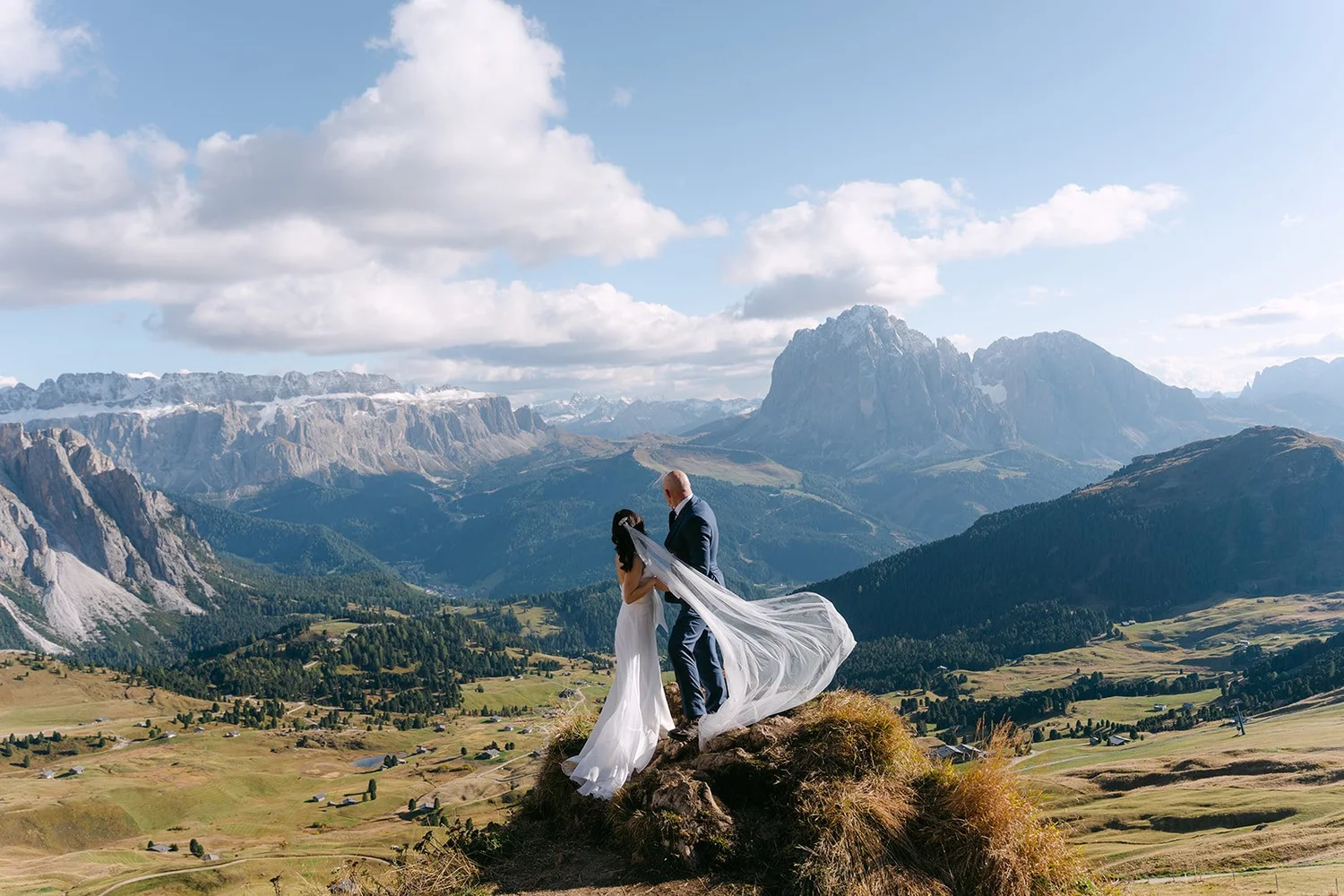 A bride and groom standing on a rock overlooking a mountainous landscape with green valleys and rugged peaks, under a sky with scattered clouds.