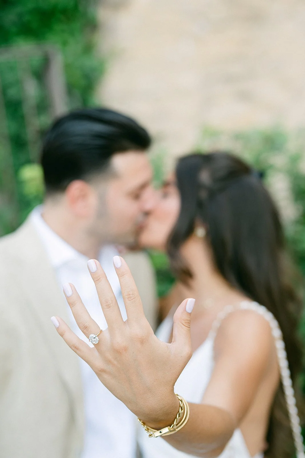Close-up of a woman's hand with an engagement ring, showing a couple kissing in the background.