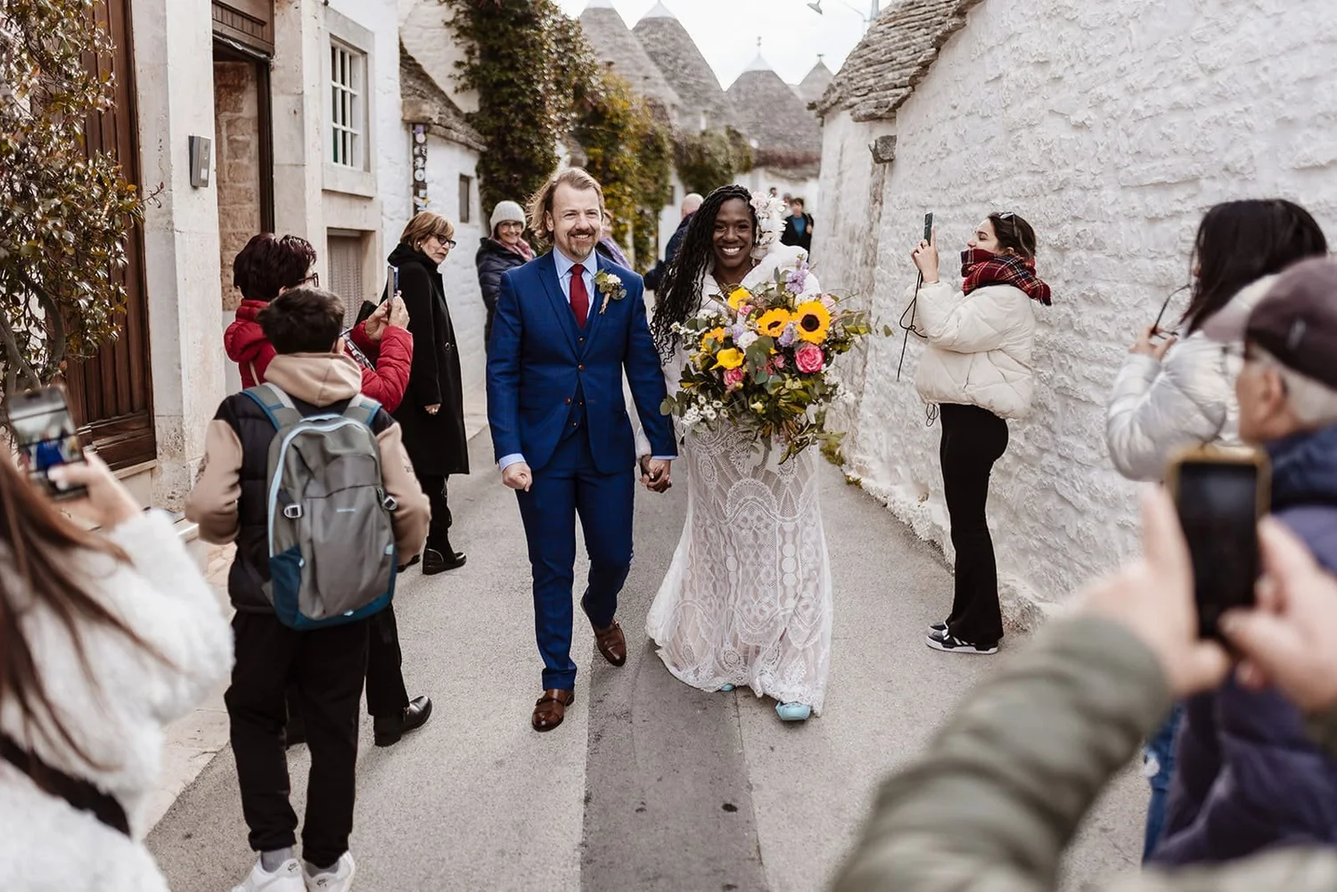A bride in a white lace dress holding a large bouquet of sunflowers and flowers, walking hand-in-hand with a groom in a blue suit, surrounded by people taking photos outside on a narrow street.