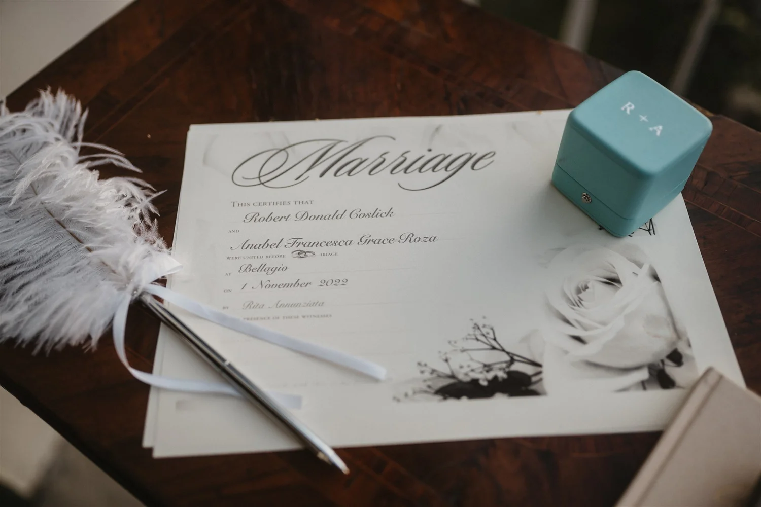 Wedding certificate titled 'Marriage' on a wooden table with a white feathered accessory and a small blue box labeled 'R + A'. The certificate includes names Robert Donald Coslick and Anabel Francesca Grace Roza, and the date November 1, 2022.