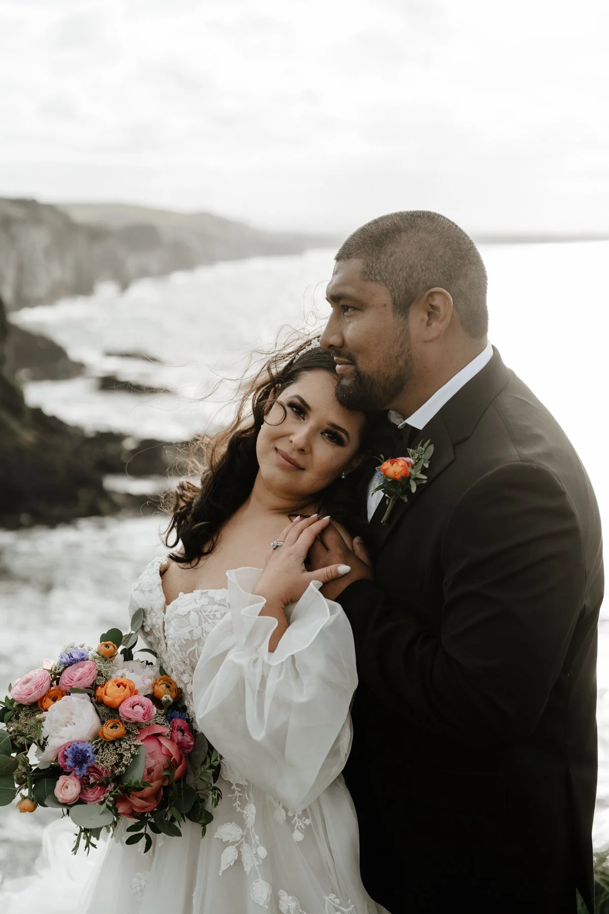 A couple embracing on a beach during their wedding, with a woman holding a colorful bouquet of flowers and the man wearing a black suit with a flower boutonniere.