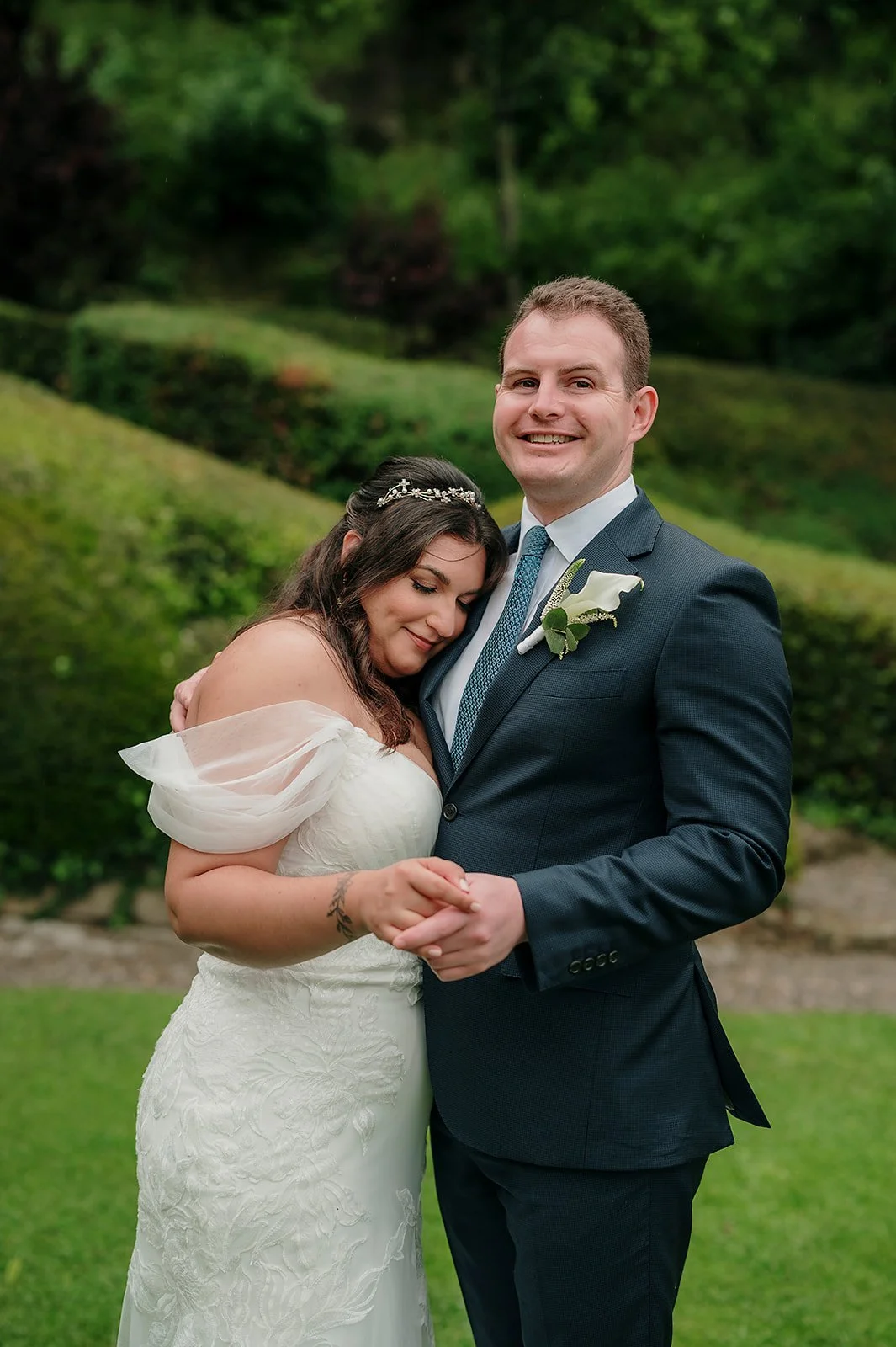 A bride and groom embrace outdoors during their wedding, with the bride resting her head on the groom's chest and both holding hands.