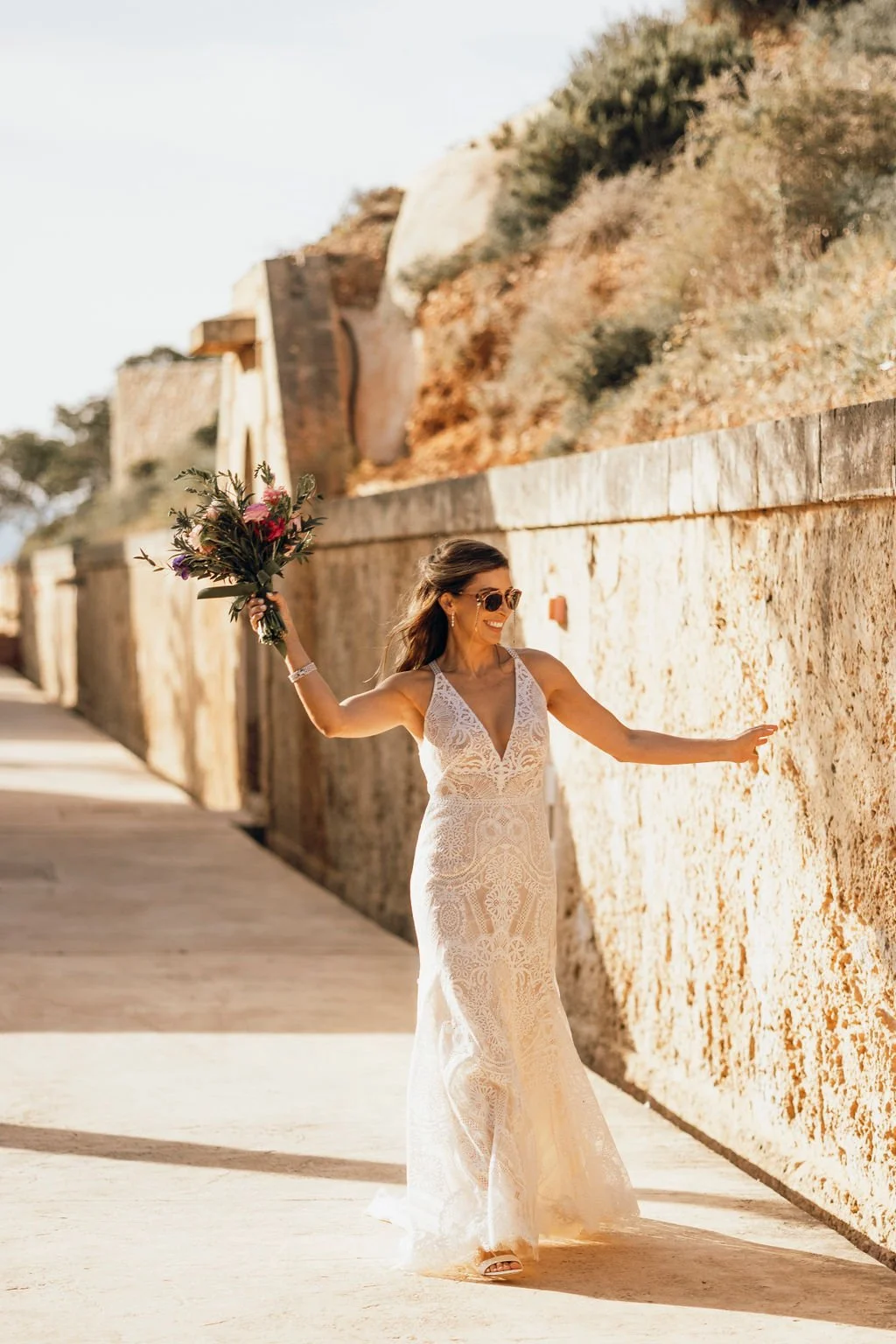 A woman in a white lace wedding dress holding a bouquet of flowers, smiling, and dancing outdoors against a stone wall with a scenic background.