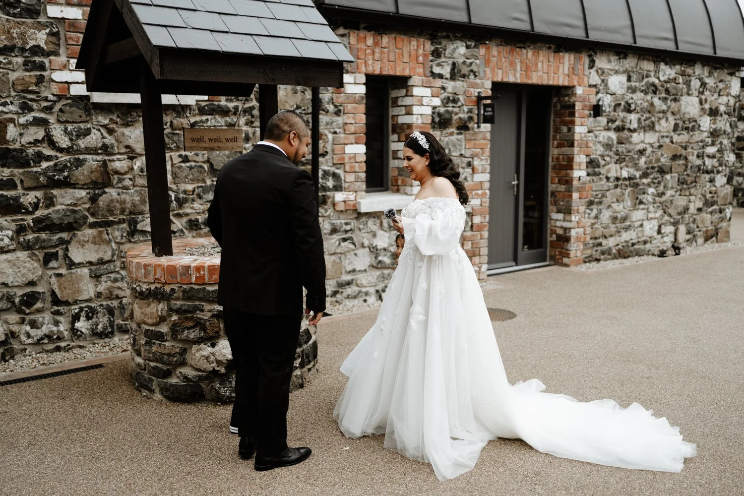 A bride in a white wedding dress and a groom in a black suit standing outdoors in front of a stone and brick wall, smiling at each other during a wedding ceremony.