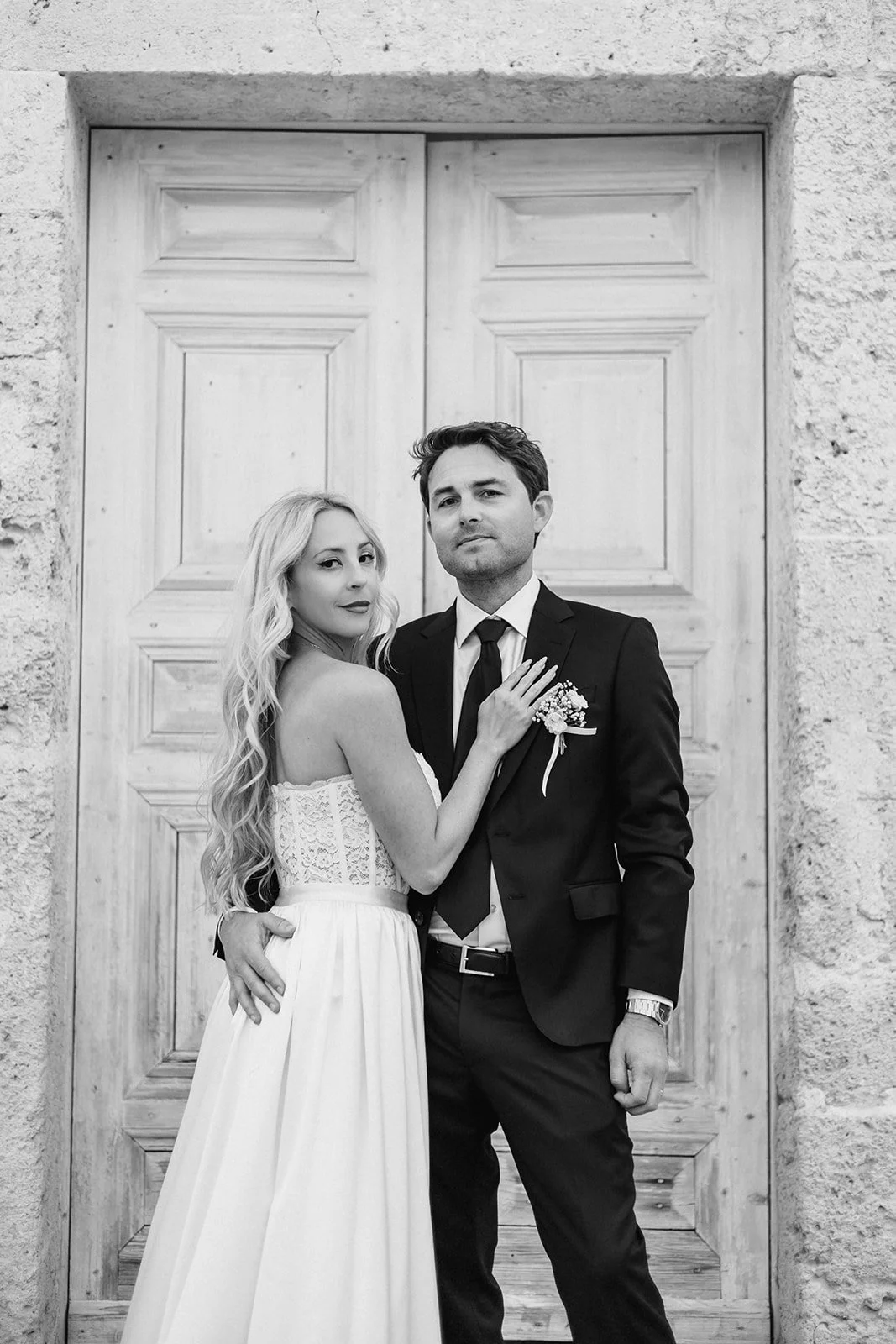 Black and white photo of a bride and groom standing together in front of a wooden door. The bride has long, wavy hair and is wearing a strapless wedding dress, while the groom is dressed in a dark suit with a tie and a boutonniere.