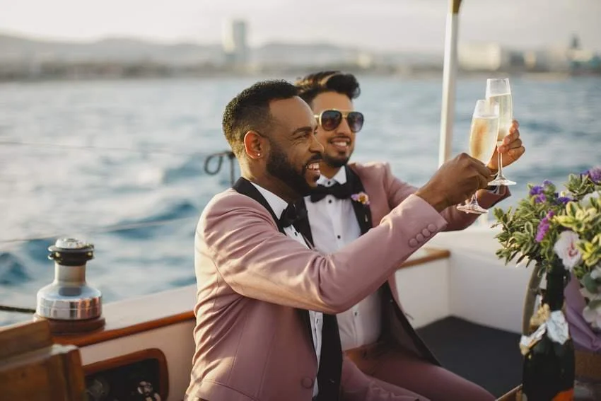 Two men in pink suits celebrating with champagne on a boat with a city skyline in the background.