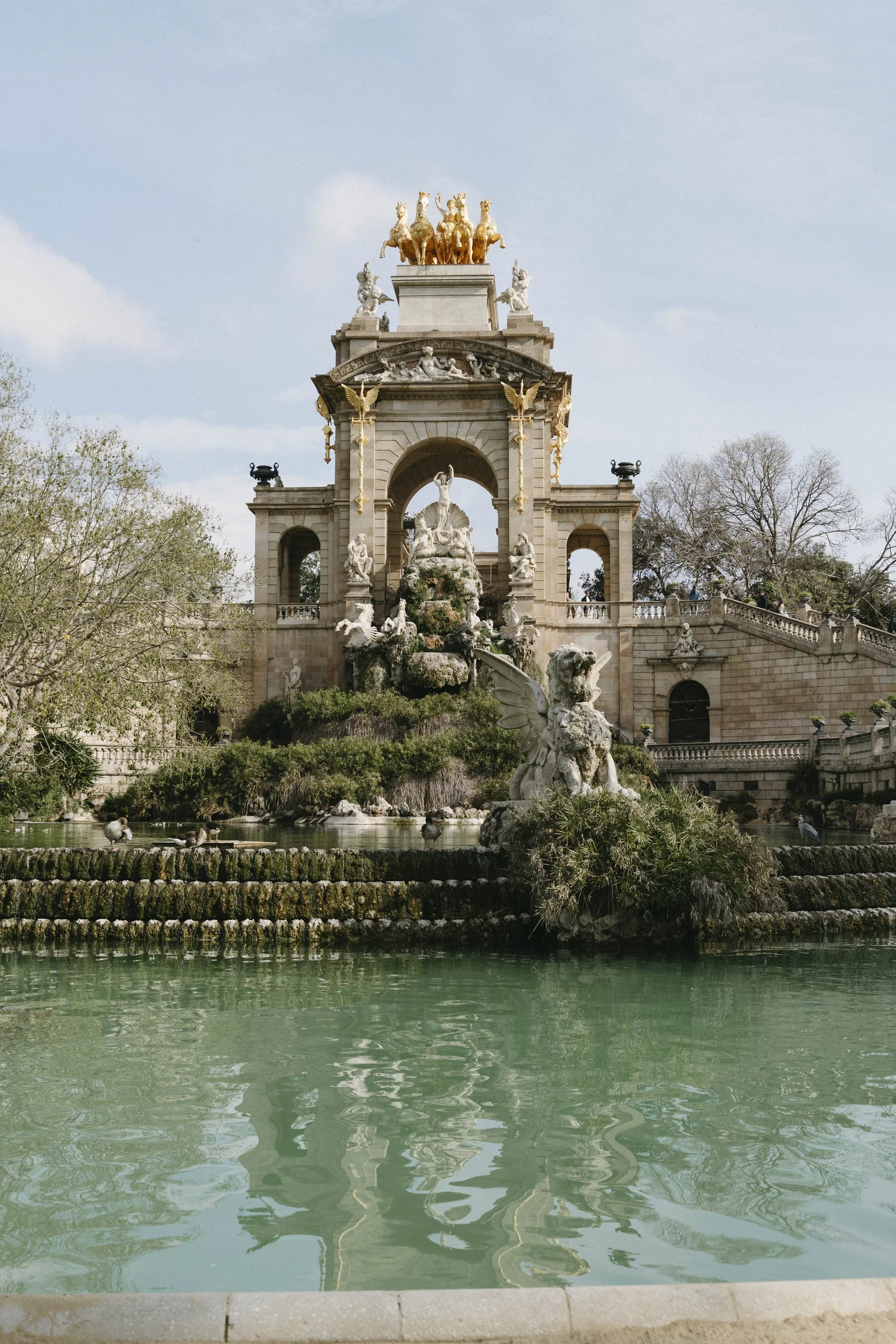 Statues and a fountain with water, decorative architecture, trees, and ducks on a pond.