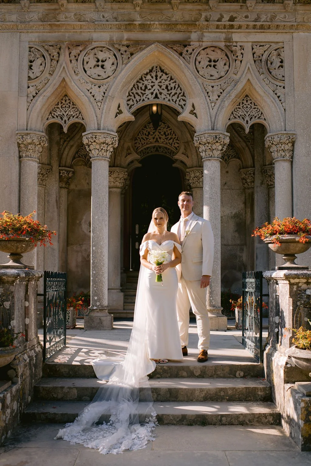 A bride and groom standing on the steps of a historic church with intricate gothic architecture, flowers, and iron railings.