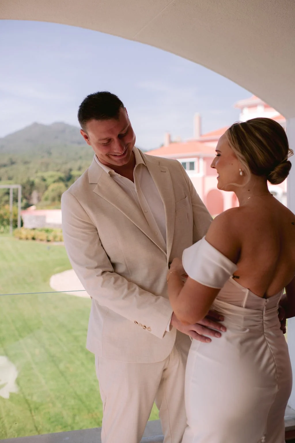 A couple dressed in formal wedding attire holding hands and smiling at each other outdoors with a green lawn and distant hills in the background.