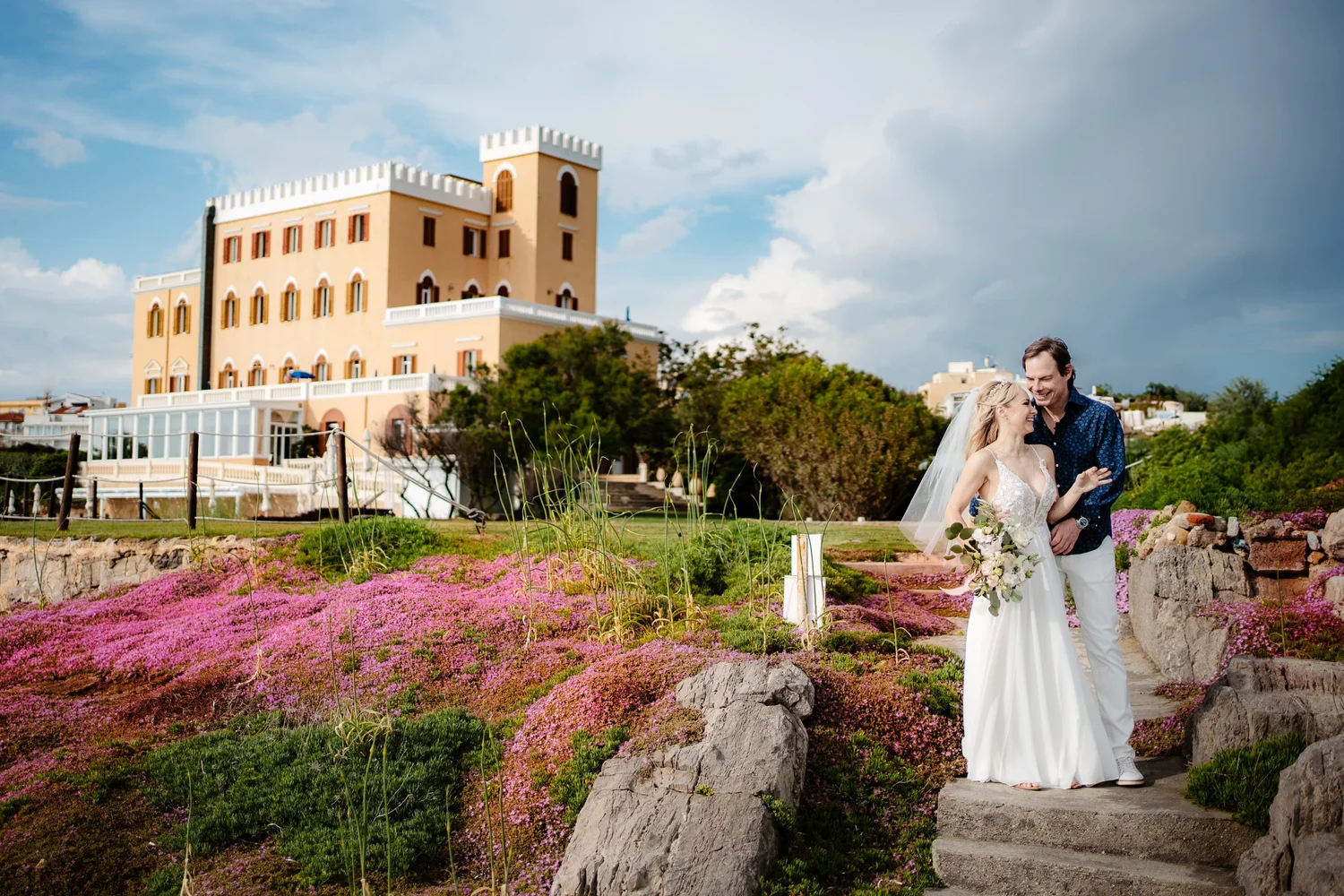 A bride and groom smiling and walking together in a garden with pink flowers, rocks, and stairs, with a large yellow castle-like building in the background under a partly cloudy sky.