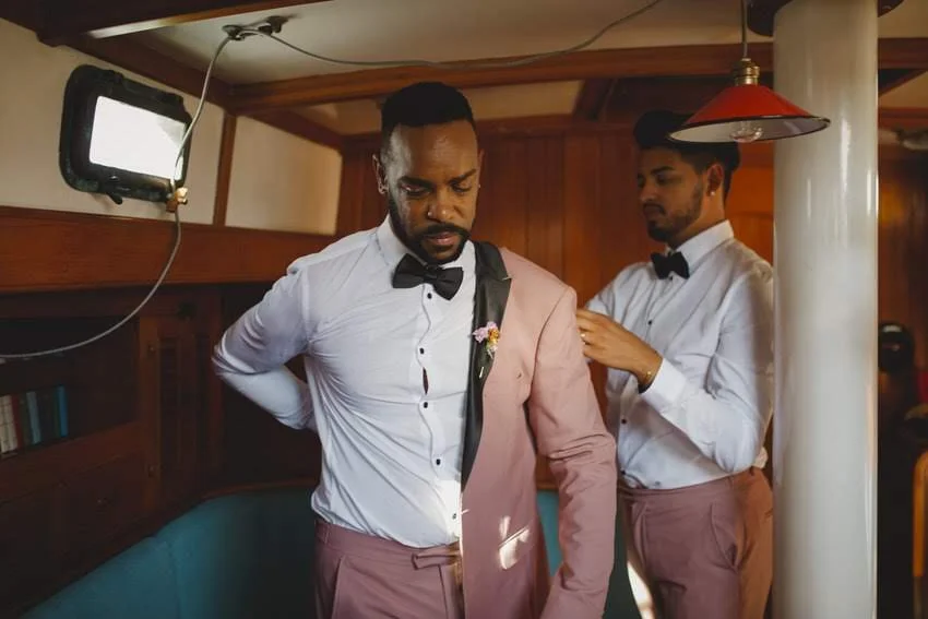Two men dressed in formal attire, with bow ties, preparing for an event. One is adjusting his tuxedo, and the other stands nearby helping. They are in a room with wooden paneling and a small wall-mounted television.