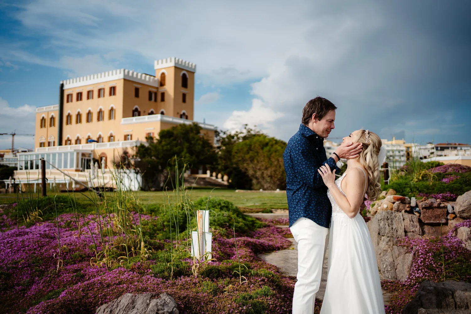 A couple dressed in wedding attire sharing a romantic moment outdoors, with a castle-like building and a landscaped garden with purple flowers in the background.