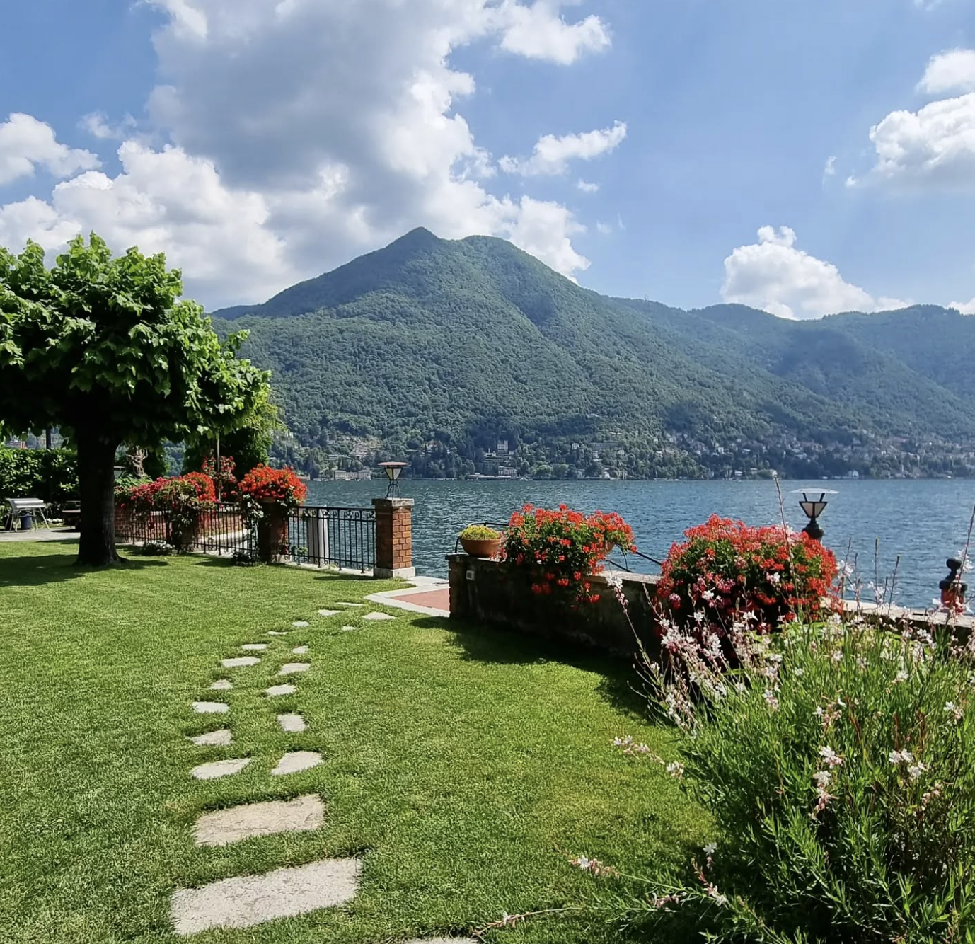 A scenic lakeside view with a grassy area, stone pathway, colorful flowers, and a view of a mountain in the background under a partly cloudy sky.