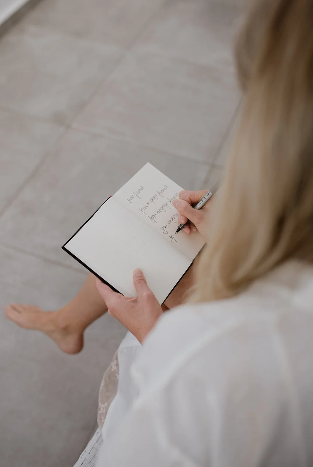 A woman with blonde hair, dressed in white, sitting on a light-colored tiled floor, writing in a small notebook with cursive handwriting.