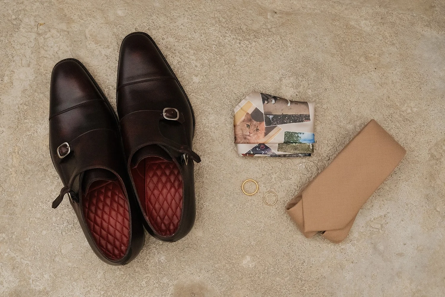 A pair of black leather dress shoes with red quilted insoles, a small folded fabric with a cat print, a rolled-up tan cloth, and two rings on a beige textured surface.