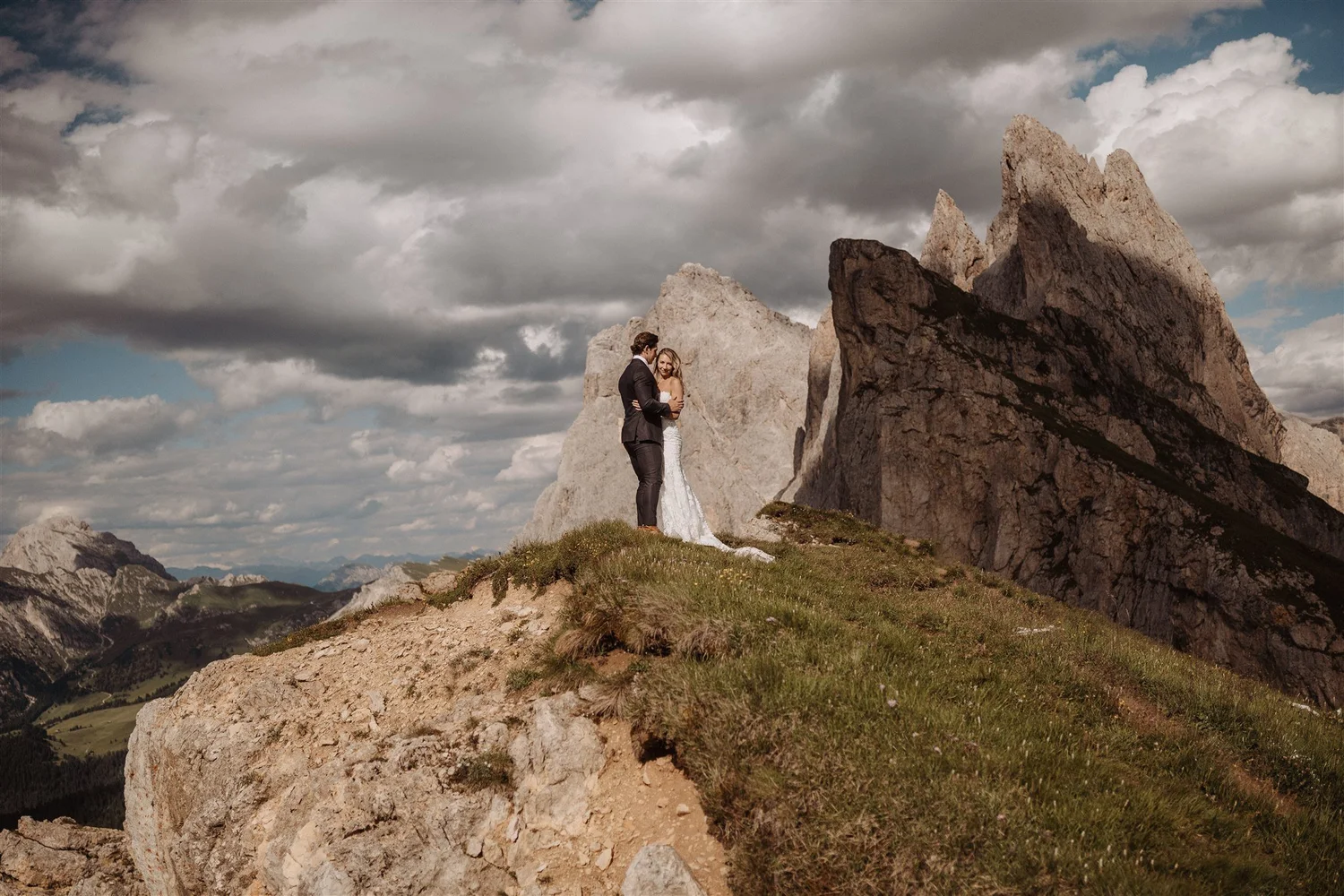 A newlywed couple standing on a grassy hill among large rock formations in a mountainous landscape under a cloudy sky.
