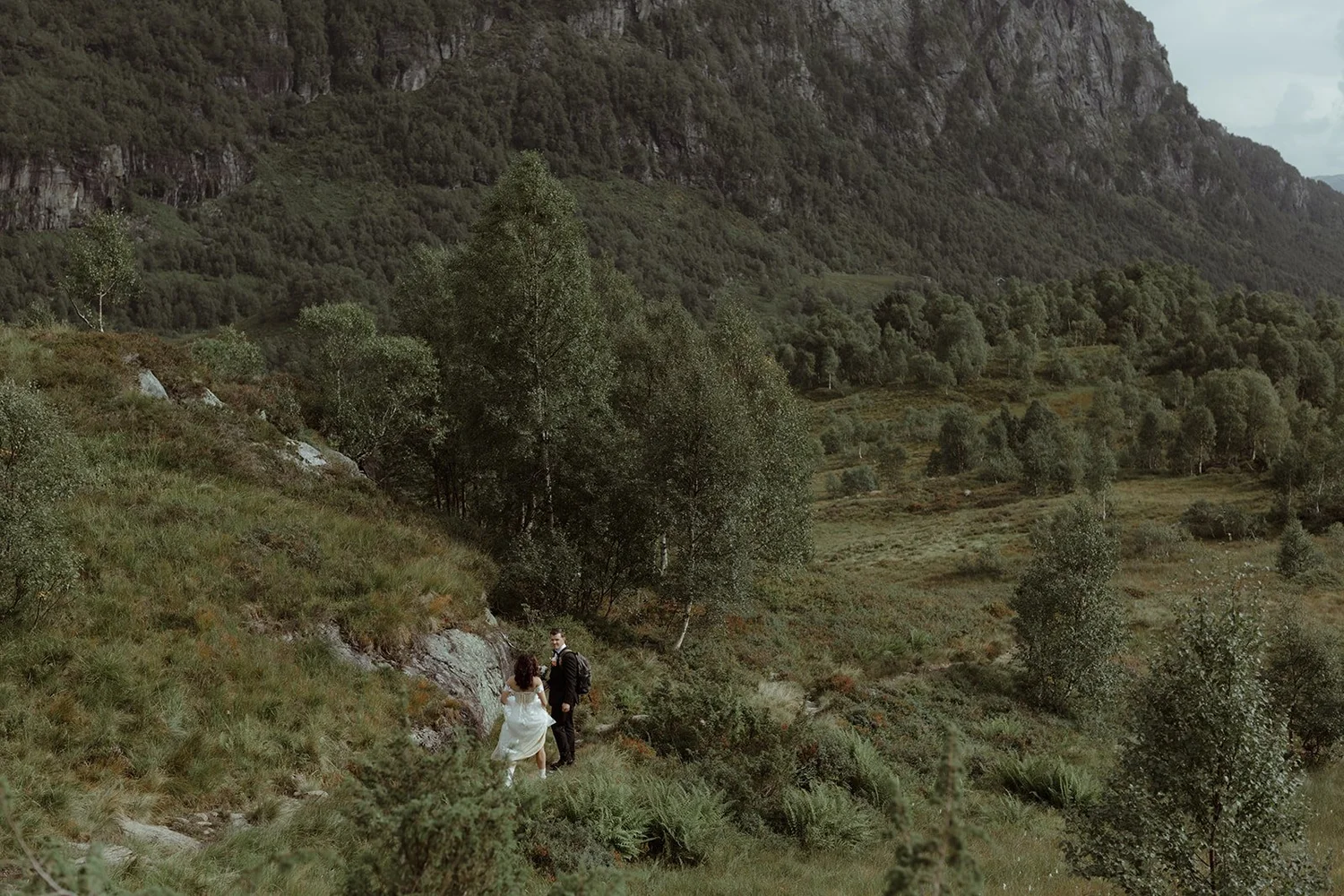 A couple, possibly newlyweds, walking through a mountainous, green landscape with trees and rocks, under a cloudy sky.