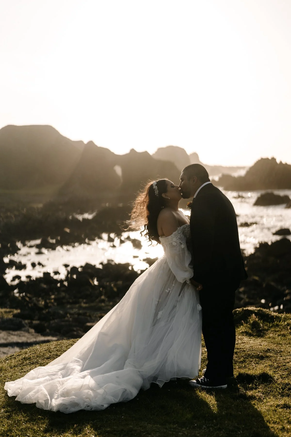 A bride and groom kiss on a rocky beach at sunset. The bride wears a white wedding dress with a long train, and the groom wears a black suit. The background features large rock formations and water reflecting the sunlight.