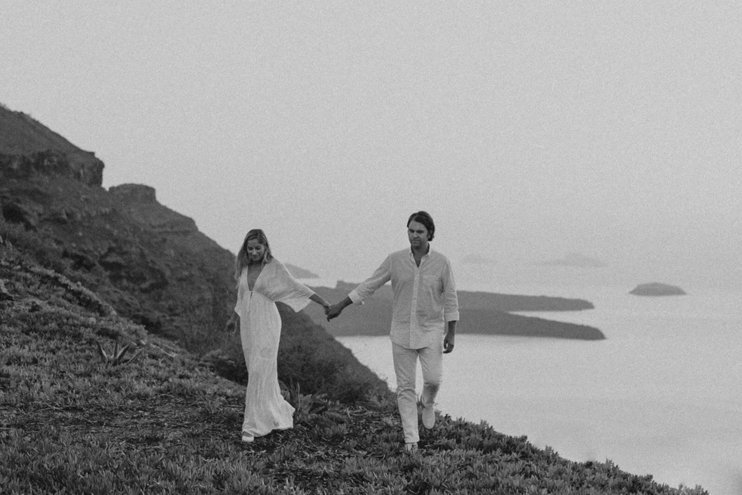 A couple walking hand in hand along a hillside coast in black and white, with mountains and water in the background.