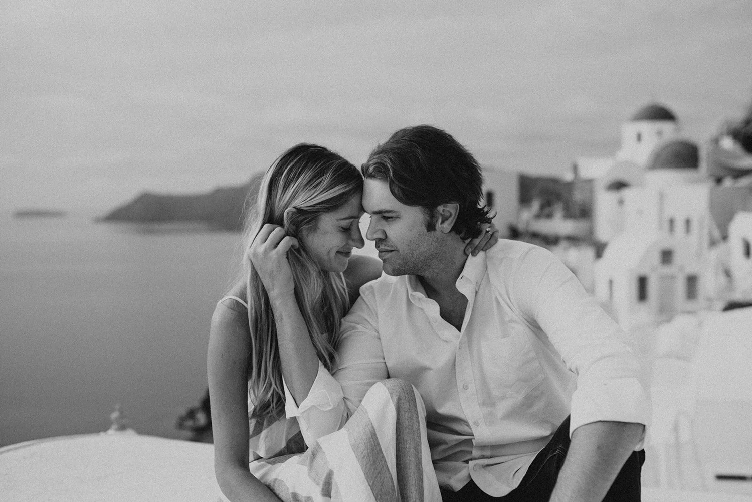 A black and white photo of a couple sitting close together with their foreheads touching, smiling softly, overlooking a coastal town with white buildings and a mountain in the background.