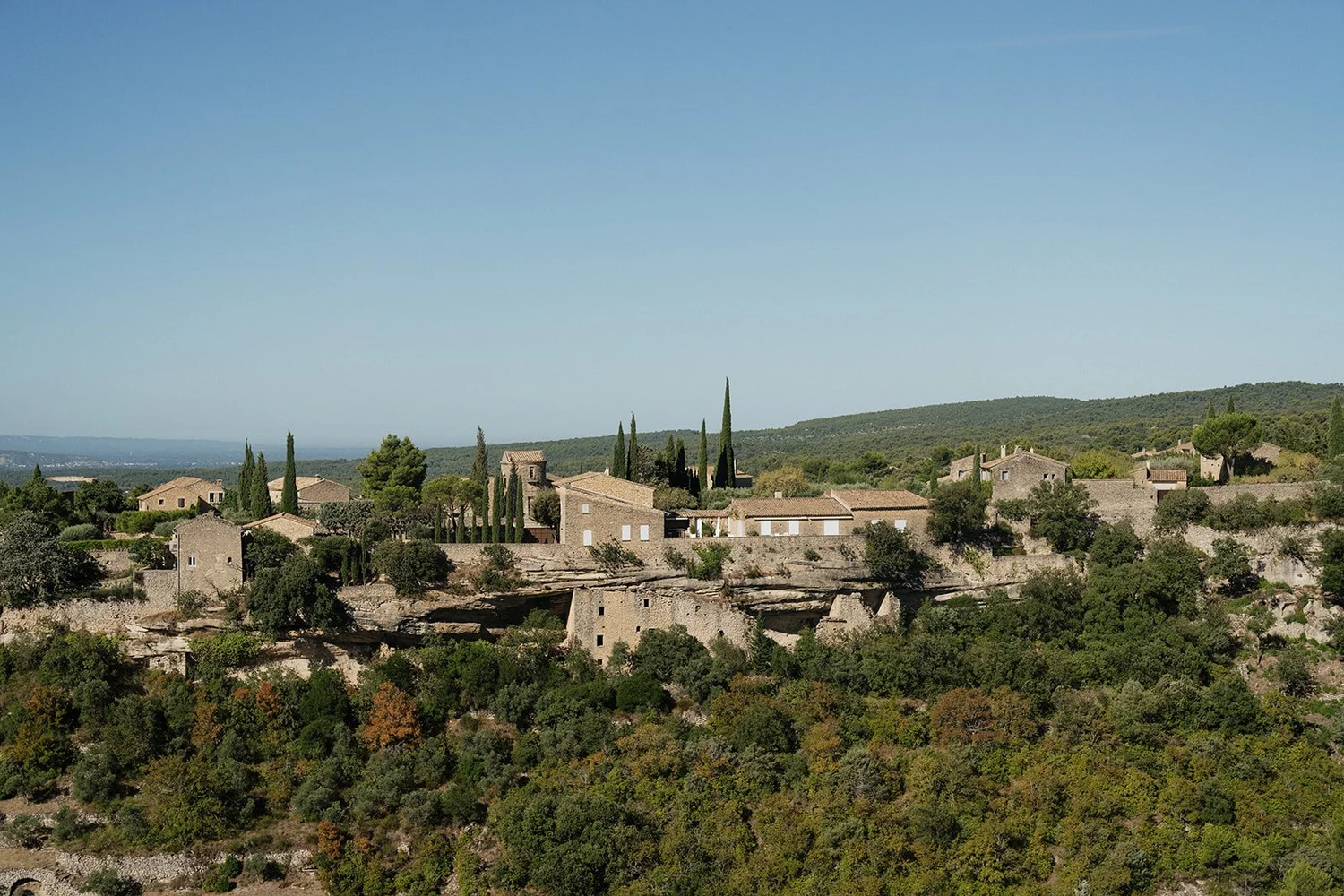 Scenic view of a hillside village with stone buildings and cypress trees, surrounded by green forest under a clear blue sky.