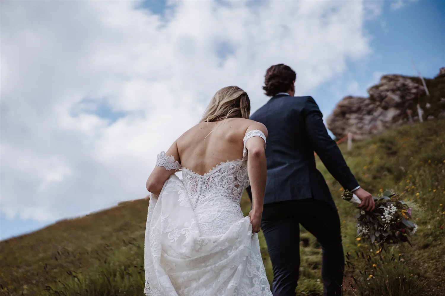 A bride and a groom walking outdoors on a hillside, with the bride holding a bouquet of flowers, seen from behind.