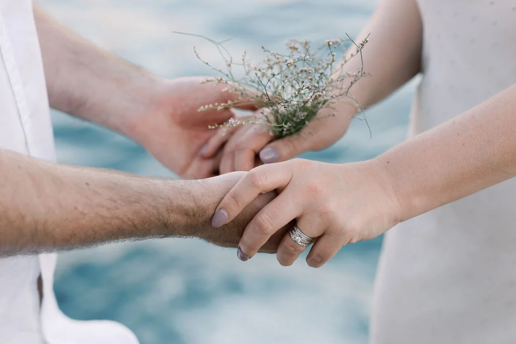 A couple holding hands during a wedding ceremony by the water, with one person placing a ring on the other’s finger and holding a small bouquet.