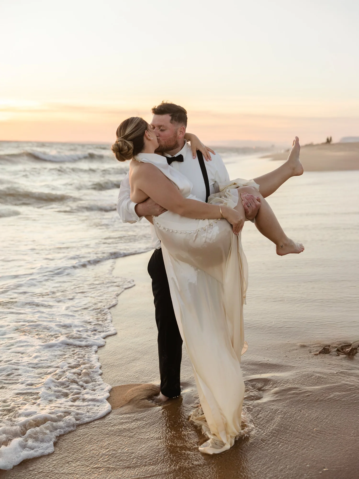 A couple in wedding attire kissing on the beach at sunset, with the groom holding the bride as she lifts one leg.